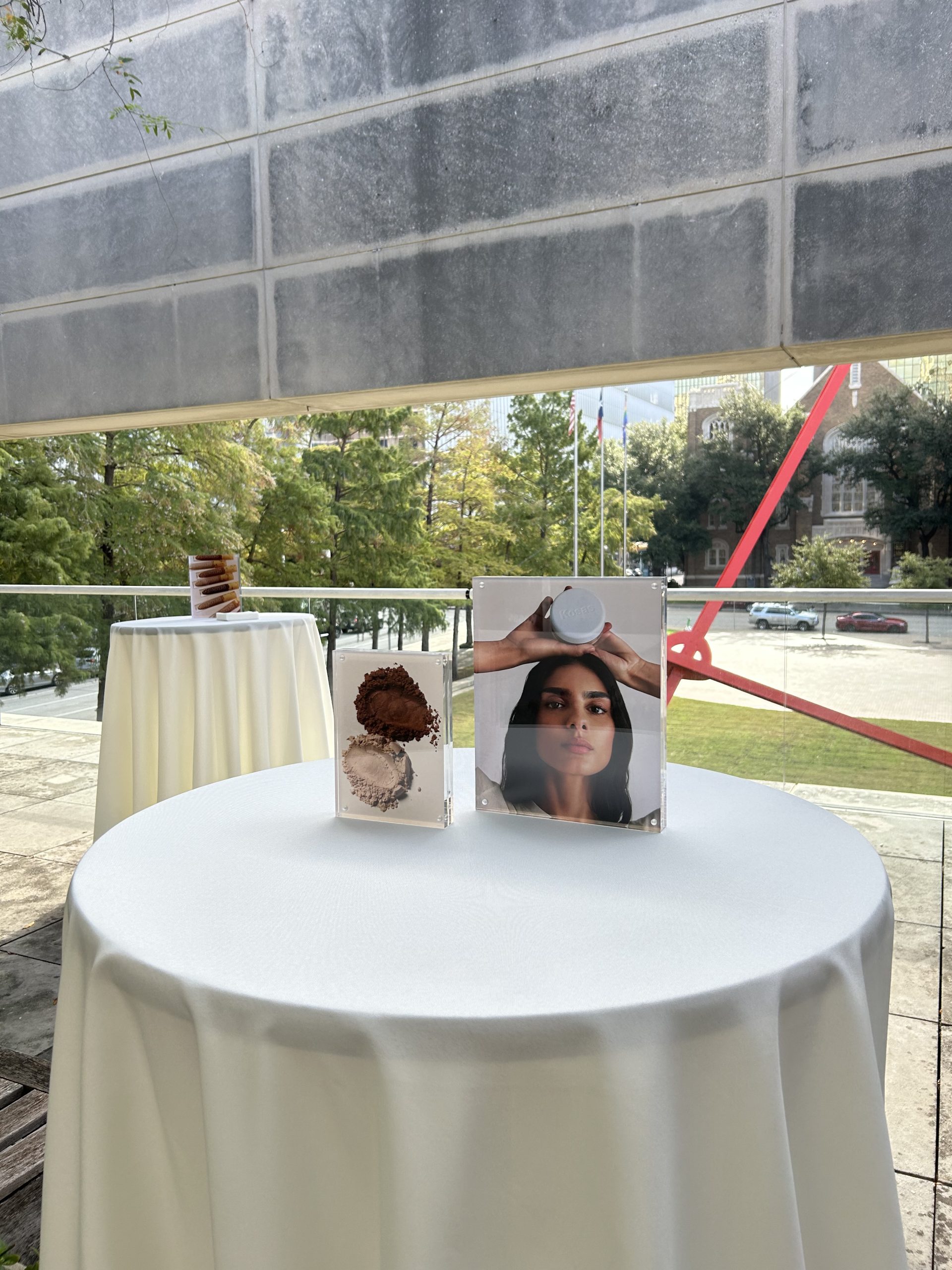 Kosas campaign cards in clear holders on a white terrace table overlooking a red sculpture.